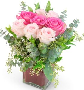 pink roses, a red ti leaf, eucalyptus, seeded eucalyptus, and myrtle professionally hand-arranged in a red cube vase, this flowery gift of love and romance for Valentine’s Day or an anniversary 