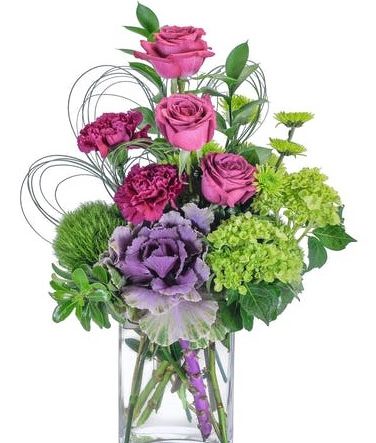 A stunning mix of magenta and green flowers, including roses, hydrangea, hypericum berry and ornamental kale, designed in a clear glass vase. The loops of bear grass shelter, and add extra interest and a delicate touch to the beautiful blooms. Purple roses, green hydrangea, and ornamental kale are designed in a clear glass vase and accented with premium foliage.