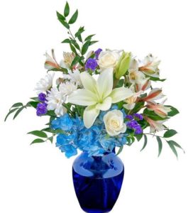 A combination of blue hydrangeas, white alstroemeria, white roses, white lilies, purple statice, and white daisies in a beautiful blue glass vase.