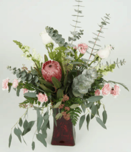 This wonderful arrangement has mink protea, pink roses, snapdragons and mini carnations accented with fun and wispy greens, while being accent by a glass red vase.